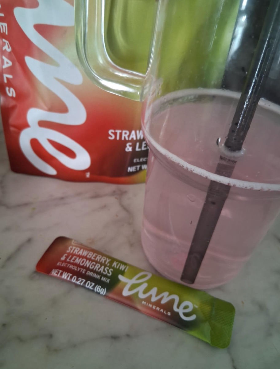 Customer photo: a pink glass of Strawberry, Kiwi & Lemongrass with a straw, next to the stick pack and pouch